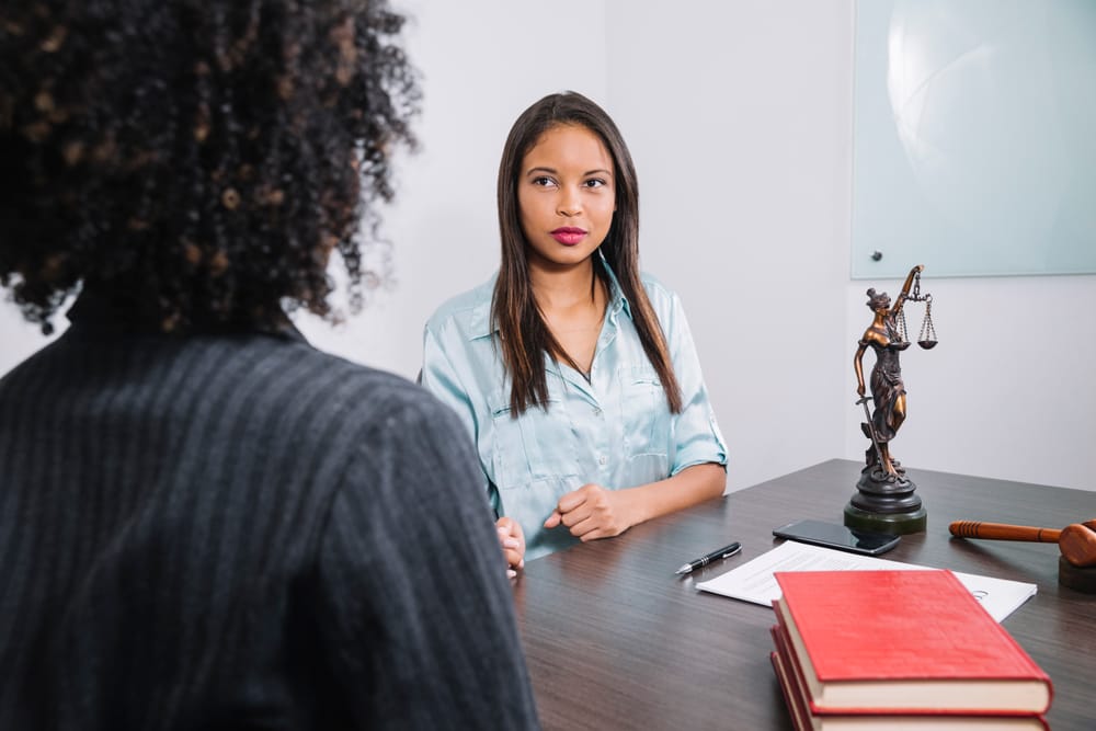 Na imagem: uma profissional de Direito (de costas para a foto) conversa com uma mulher sentada do outro lado da mesa