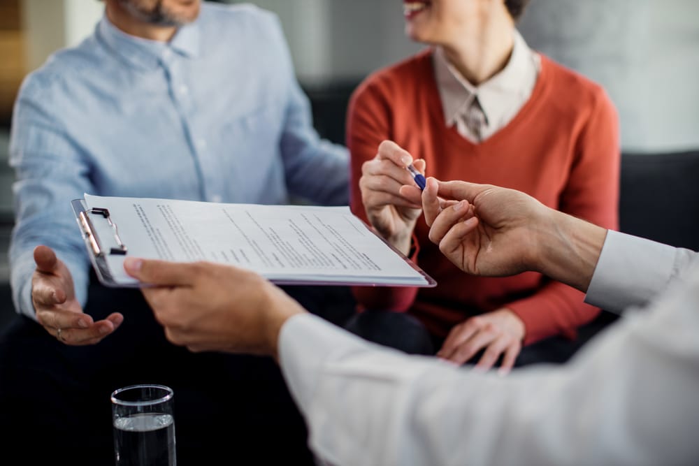 Na imagem: casal recebe documento contratual das mãos de um profissional para assinar