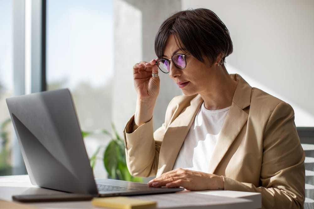Na imagem: mulher de blazer e óculos olha com atenção para a tela do notebook