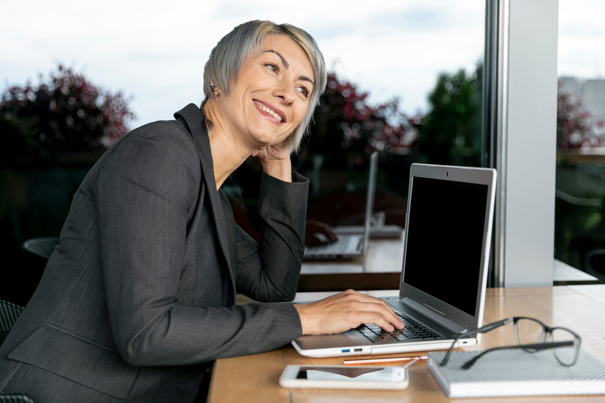 Na imagem: advogada de cabelos grisalhos parece feliz e tranquila sentada numa mesa com o seu notebook