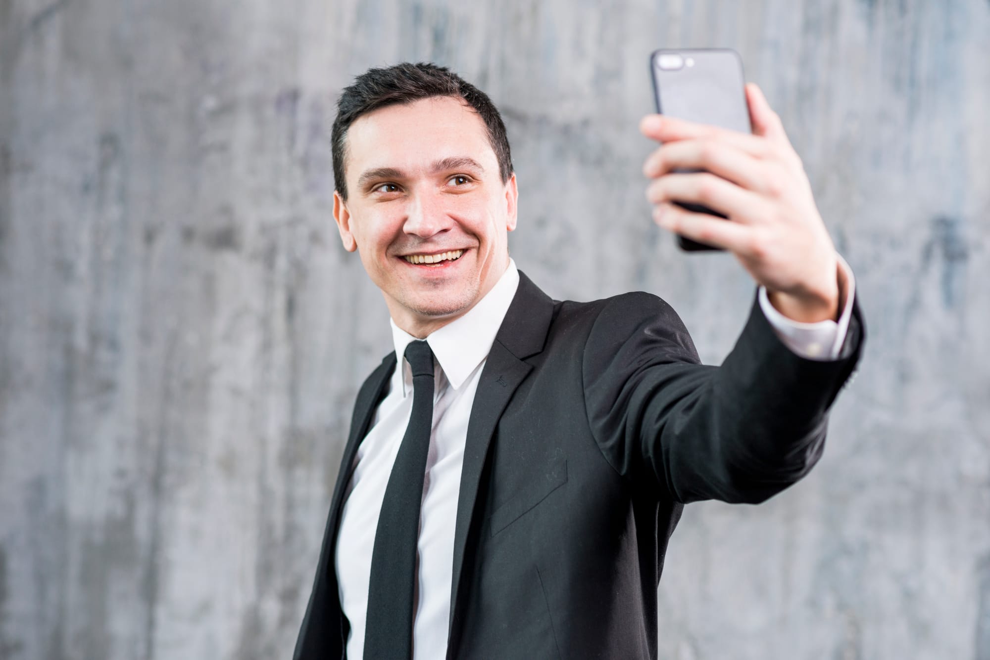 Na imagem: homem de terno e gravata sorri para câmera de selfie do celular.