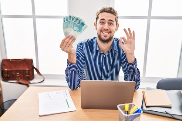 Na foto: Homem atrás de uma mesa de escritório levanta notas de 100 reais e sorrindo