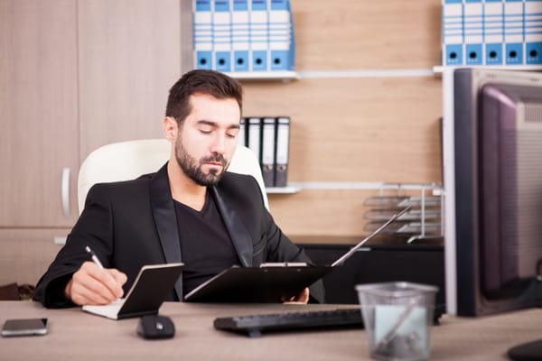 Na imagem, homem está em um escritório, em frente ao computador, analisando pastas, anotando dados com olhar analítico