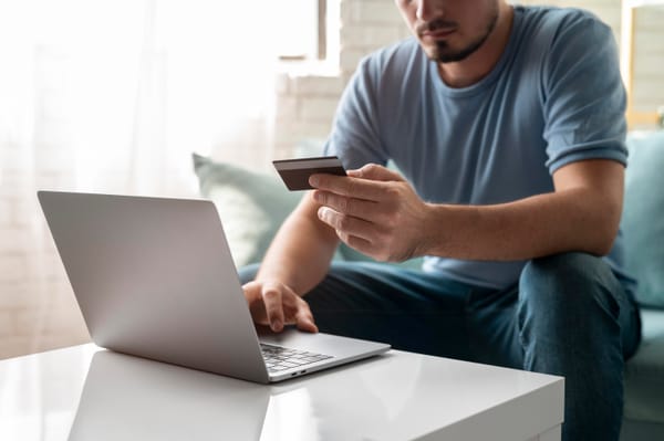 Na imagem: Homem está sentado em sofá, segurando o cartão de crédito e digitando no teclado do notebook.